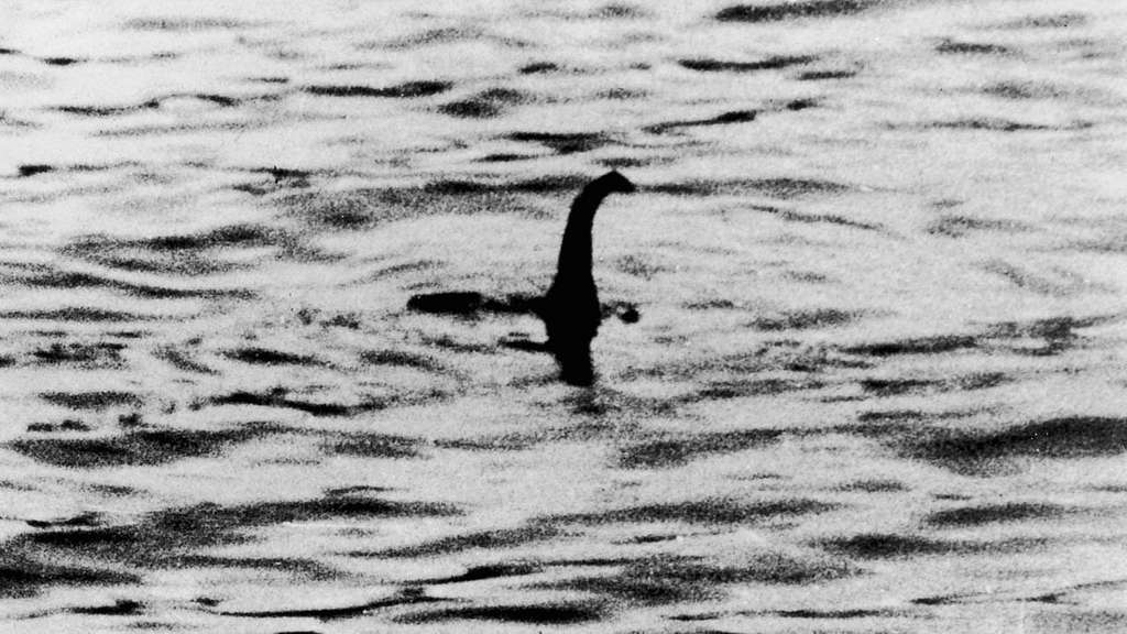 A black and white still of a creature with a long neck and small head sticking up out of rippling water.