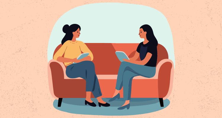 Two women sitting on a couch with tablets/books in their hands talking.