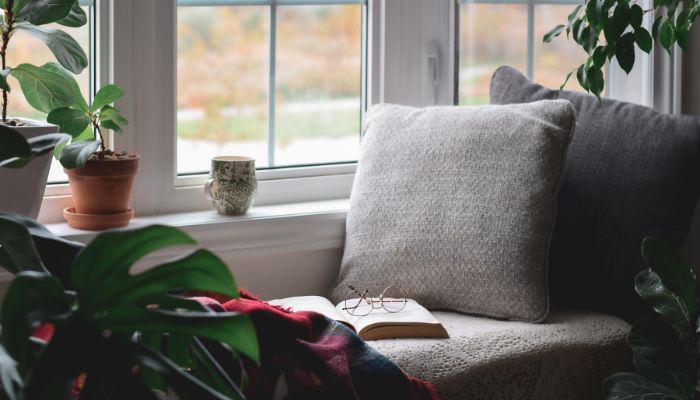 A plush seat near the window with a pillow and plants. A blanket, an open book, and reading glasses sit on the plush seat. A cup sits on the windowsill next a potted plant.