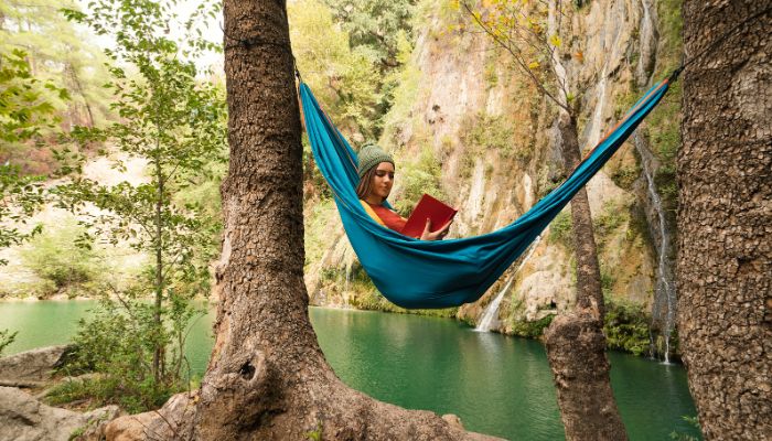 A young woman sits in a blue hammock, reading. A green lake, trees, and mountain are in the background.