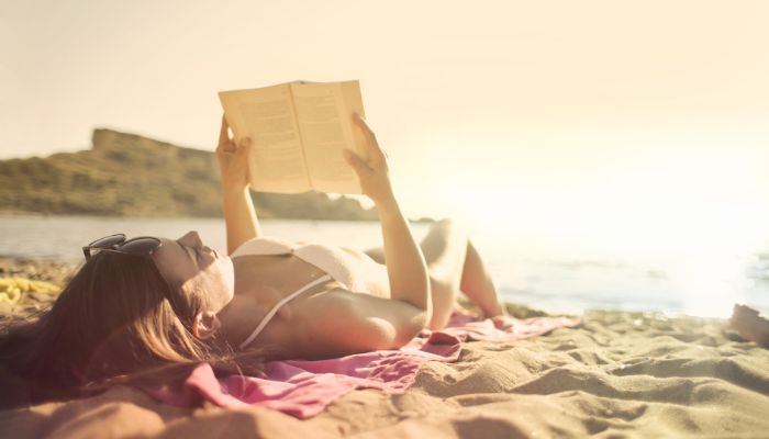 A woman lying on a beach towel reading on a sandy beach with the water and small mountain in the background.
