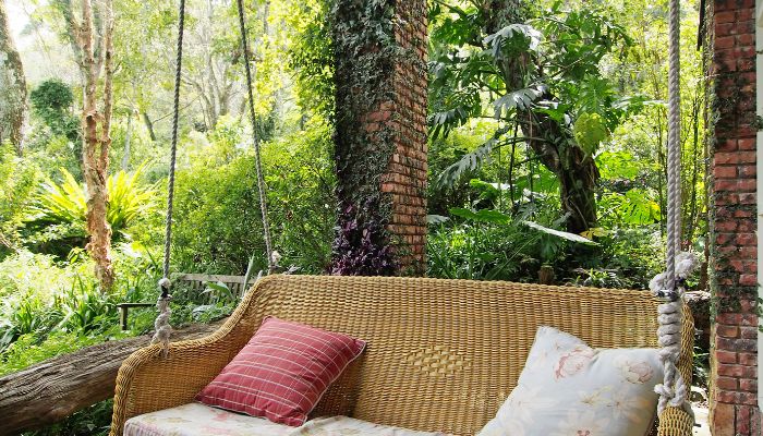 A porch made of brick with a porch swing with pillows on it. Behind the porch is a luscious forest of trees and grass.