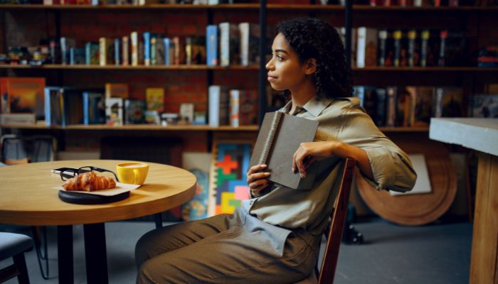 A woman sitting at a table in the library. a croissant and cup sitting on the table next to a pair of glasses. She holds a book against her while looking up. Behind her are shelves of books and children's toys.