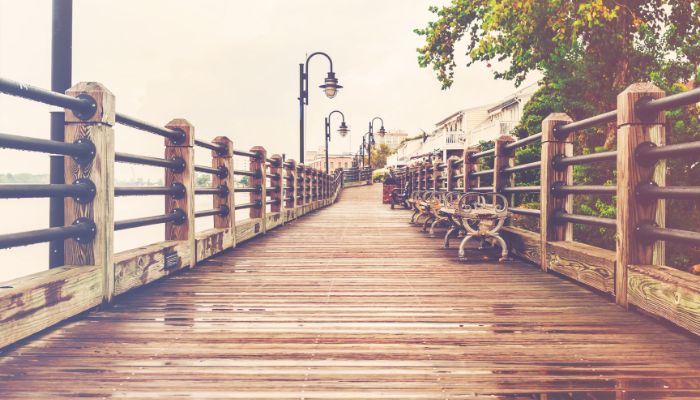A boardwalk with thick rails on each side and streetlamps hanging off the left side rails. Benches on the right side. Trees and dwellings behind the right side rails.