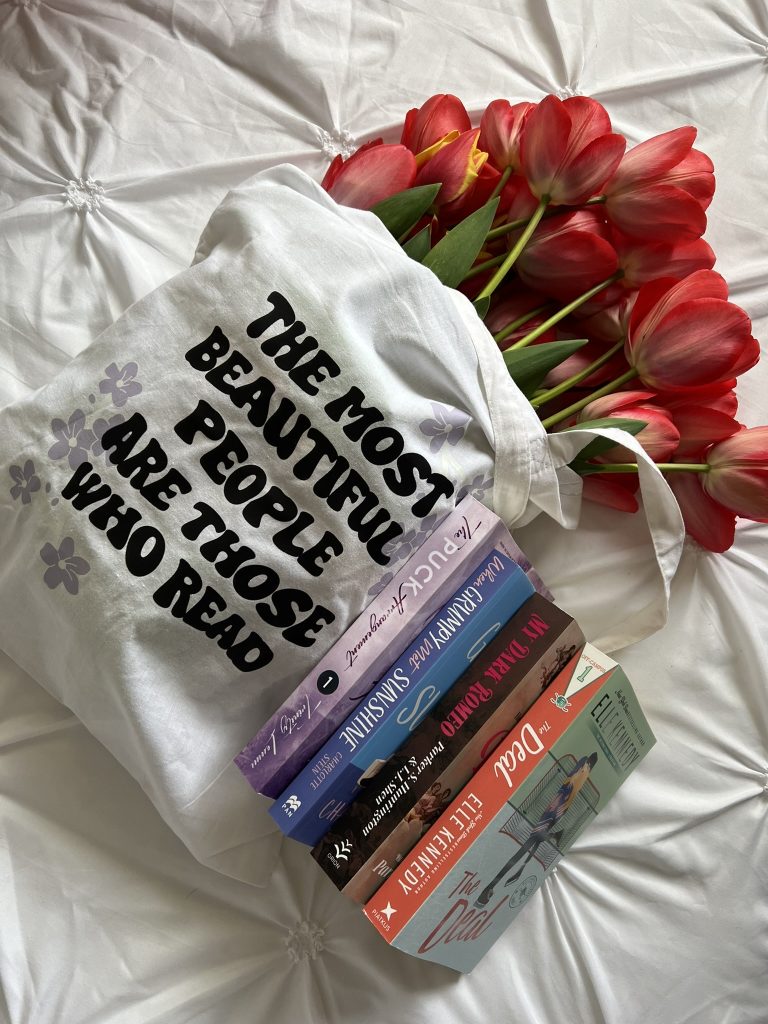A white tote bag with purple flowers and the phrase "The most beautiful people are those who read" hold a bouquet of red tulips. Four books are leaning against it, spines up. Everything is on a white fabric.