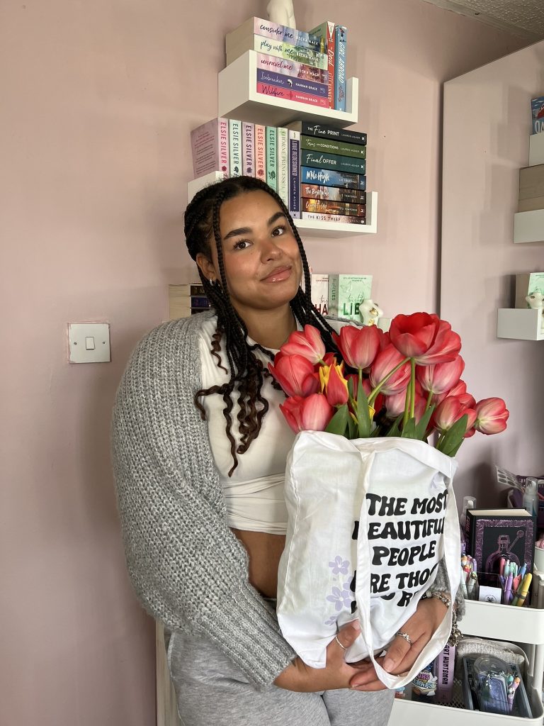 Tia of @tias.bookshelf_ poses in a gray cardigan, holding a white graphic tote bag with red tulips. She is in front of a pink wall with tiny wall shelves stacked with books.