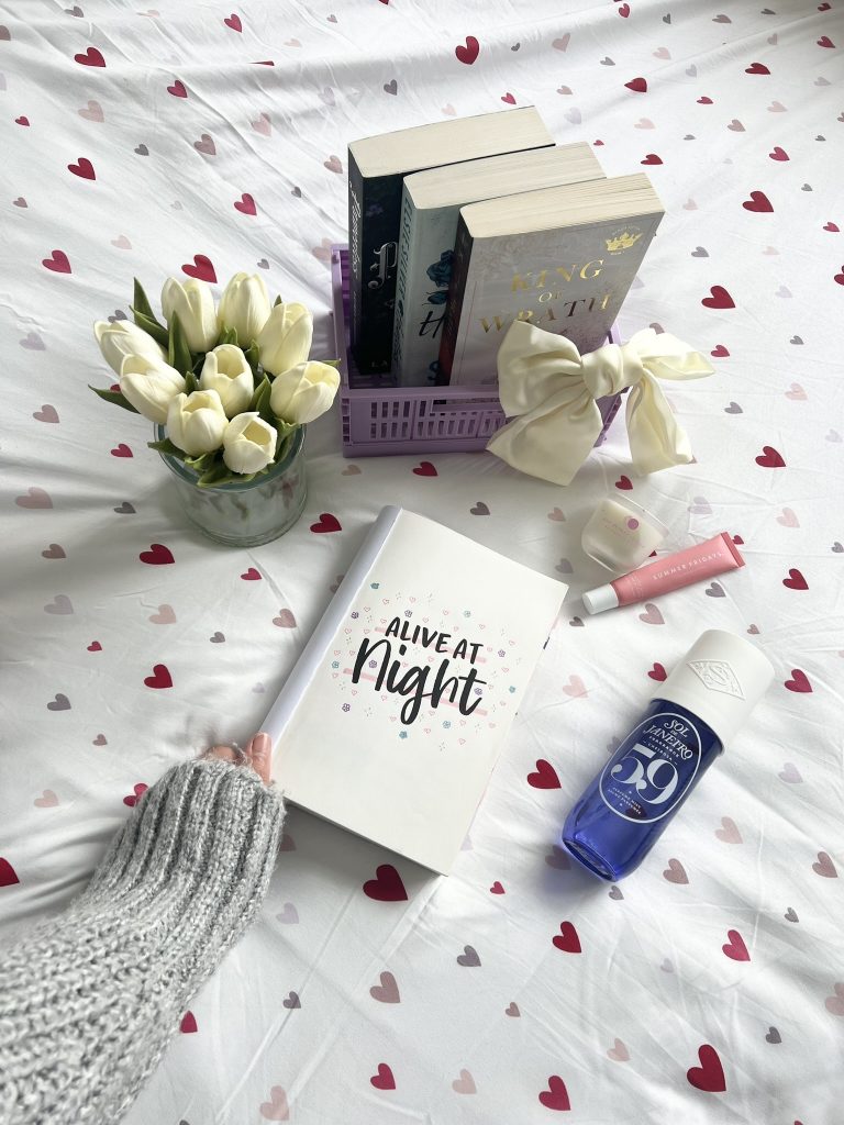 The title page for a book titled "Alive at Night" sits on a bedspread with red, pink, brown, and gray hearts. A hand with a gray sweater holds the corner of the book. A vase of white tulips, a blue perfume, a pink lipstick, a white jar of skincare, and a purple crate with three more books and a white bow frame the novel.