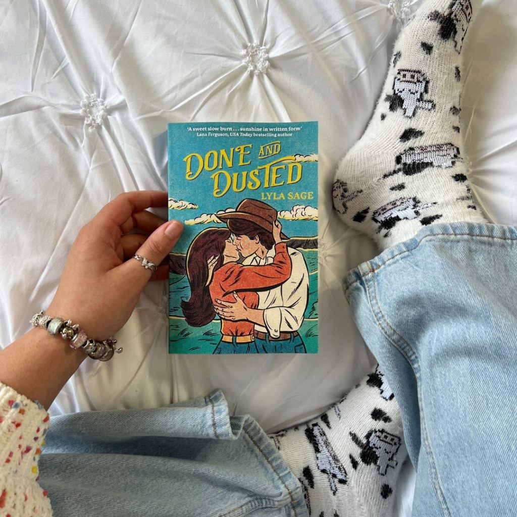 A person sitting on a white bed wearing a white sweater with multi-colored dots, blue jeans, and cow socks holds Done and Dusted by Lyla Sage in front of their crossed legs.