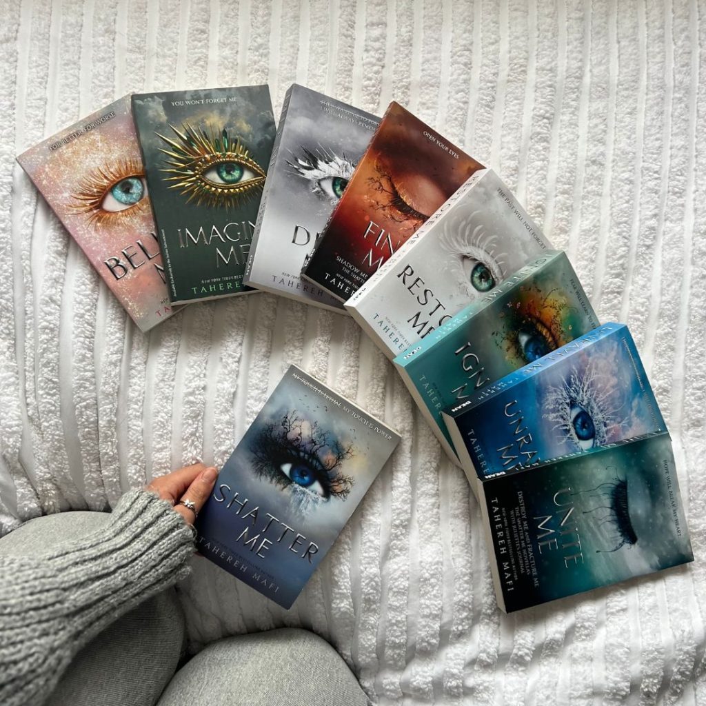 Books in the Shatter Me series are arranged in a half circle, overlapping one another on a white fabric. A hand in a gray sweater holds the first book underneath the circle.