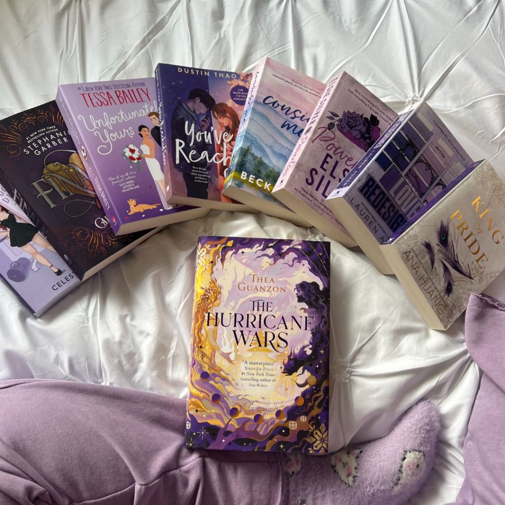 Books with covers with purple color schemes are arranged overlapping in a half circle. In the middle, on top of a pair of legs in purple socks and sweatpants, is the Hurricane Wars by Thea Guanzon.