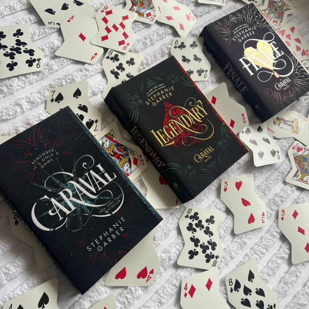 Three hardcover Stephanie Garber books are lined up, framed by wavy playing cards on a white fabric.