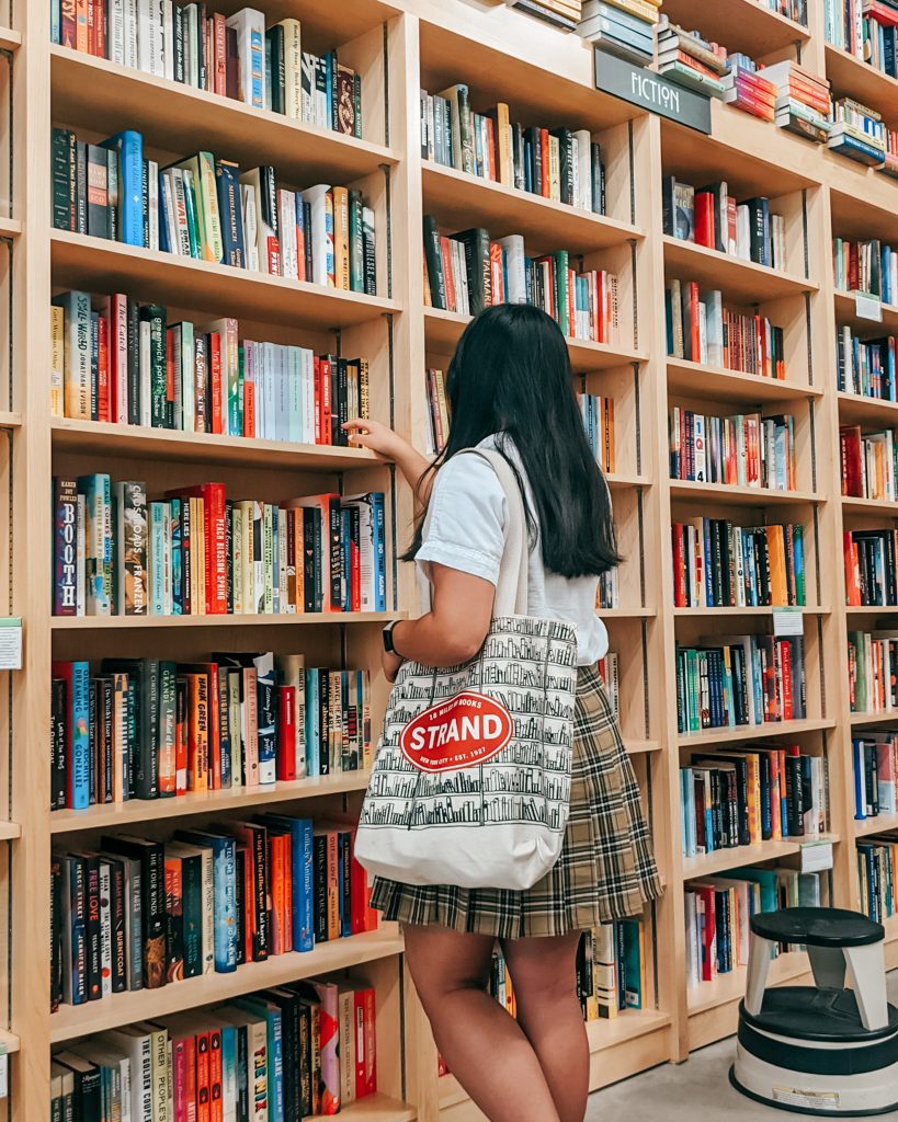 Photo of Bookstagrammer Esther of @estherfungreads reaching for a book on a bookstore's shelf. She has a tote bag from The Strand. She is wearing a white blouse and yellow plaid skirt.