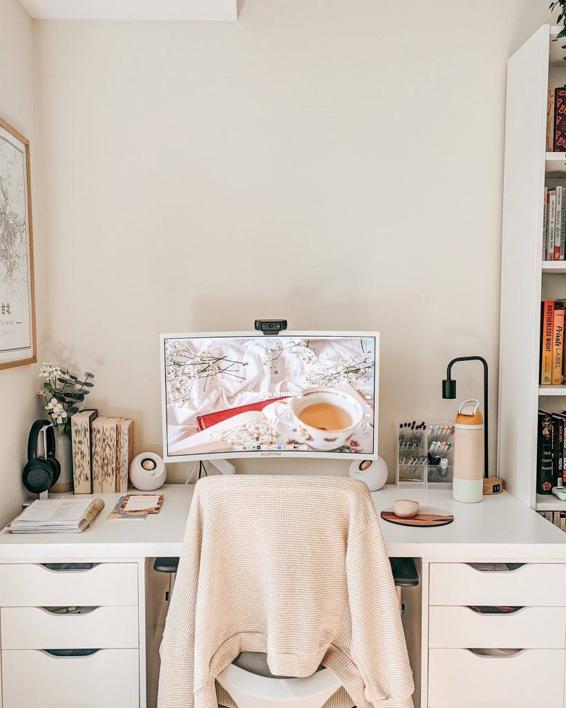 A white desk against a beige wall. A white computer monitor is placed in the center, showing an aesthetic image of a mug of tea on a bed with flower stems. Books, notebooks, pens, and speakers are organized neatly on the desk. An orange water bottle and black lamp sit on one end, and a vase of white flowers are on the other end. A framed photo hangs on the left wall, and bookshelves are on the right.