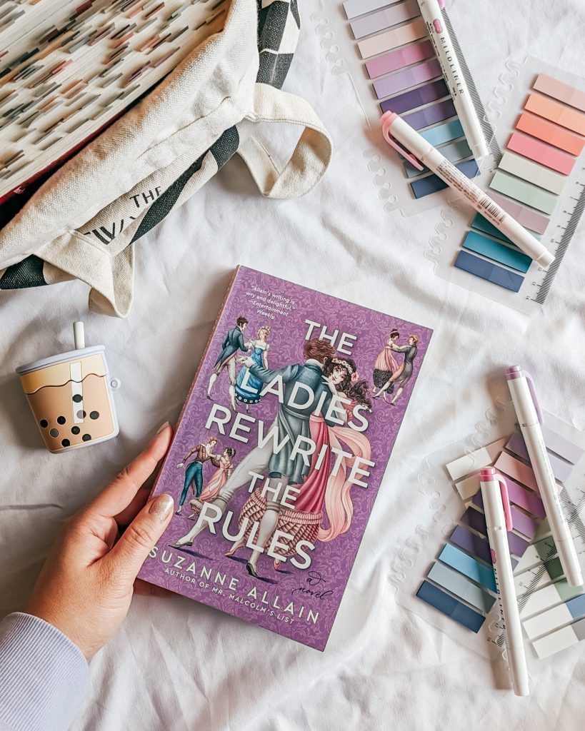 On a white cloth, a hand holds The Ladies Rewrite the Rules by Suzanne Allain. Framing the book are a tote bag, a rubber case shaped like boba tea, highlighters and annotation tabs whose colors match the purple and pink color scheme of the book.