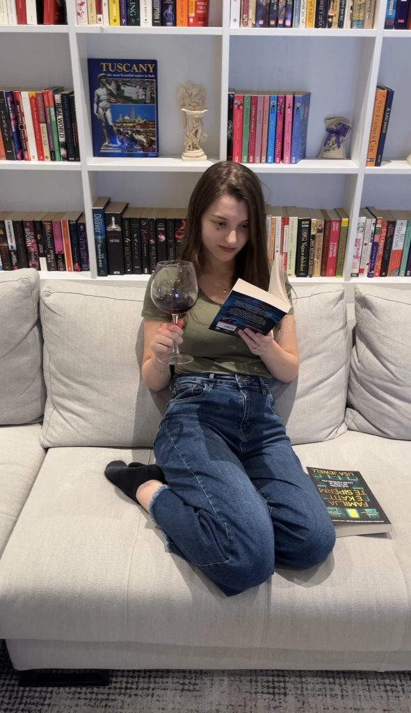 Photo of Bookstagrammer is Sara, @saras_mystery_bookclub sitting on a light grey couch with a book in one hand and a wine glass in another. Behind her are white bookshelves staked with colorful books.