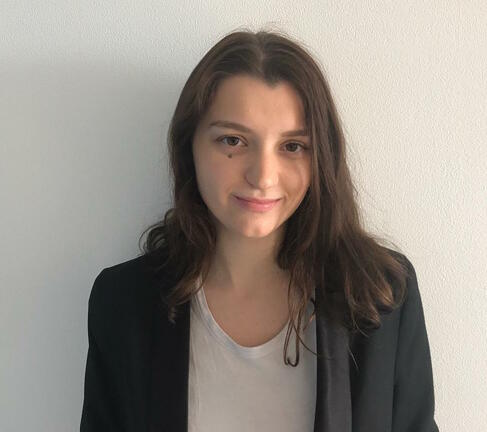 Photo of Bookstagrammer is Sara, @saras_mystery_bookclub. She stands in front of a white wall in a black blazer and white shirt. Her long brown hair is down.