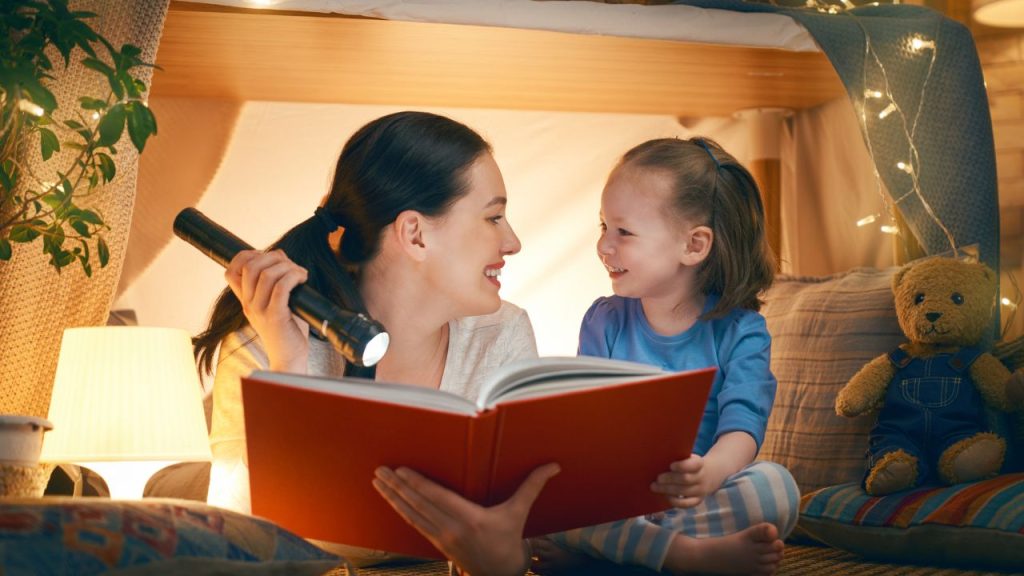 mom holding a book and flashlight, her daughter siting next to her. They're both smiling