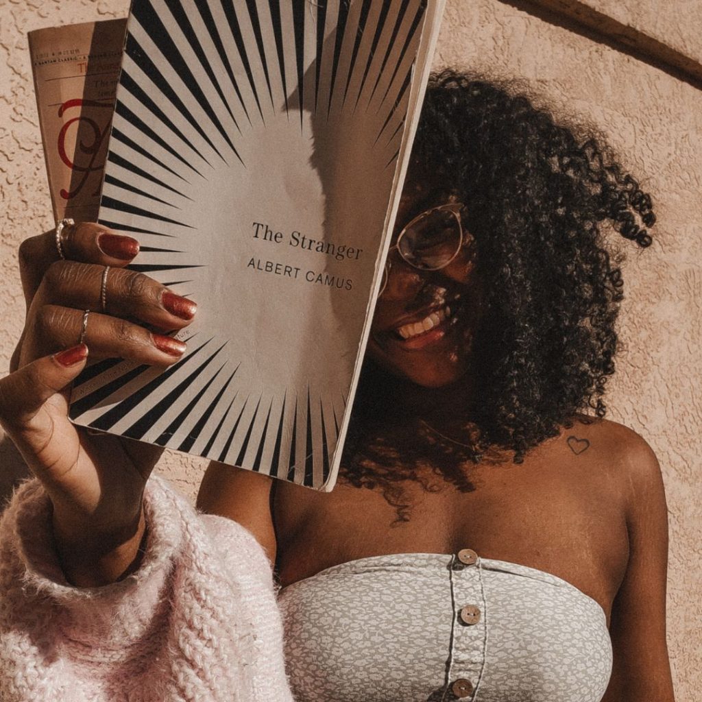 Image of Bookstagrammer Eboni, @thelovelythings505. She poses in front of a textured wall in a floral tube top and slouchy cardigan, holding up The Stranger by Albert Camus partially in front of her face.