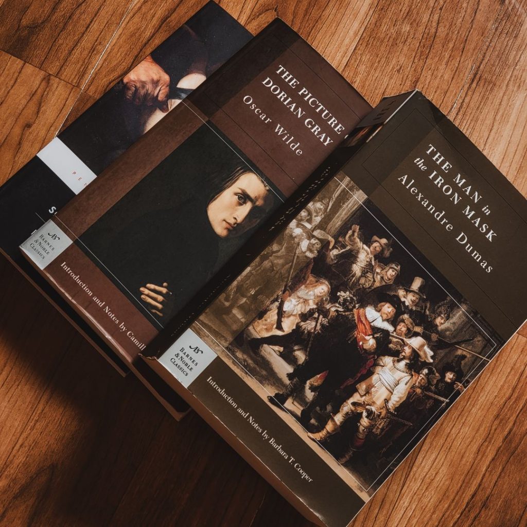 Three classic literature novels are overlapped on a wooden table. The first one is The Man in the Iron Mask by Alexandre Dumas and the second one is The Picture of Dorian Gray by Oscar Wilde. The third book on the bottom is obscured, but has a dark cover. The first book has an old painting of a group of men with guns and the second book has an old portrait of a young man.