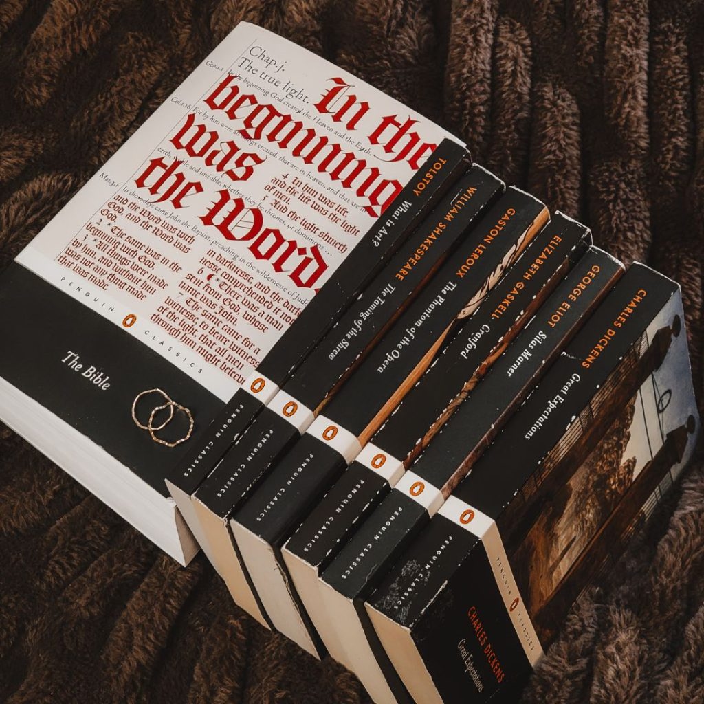 A collection of six matching Penguin Classics books are stacked spine up, with the Penguin Classics' edition of The Bible on the end, cover up. They are all placed on a dark brown blanket. Two rings are stacked on the black title strip of The Bible.