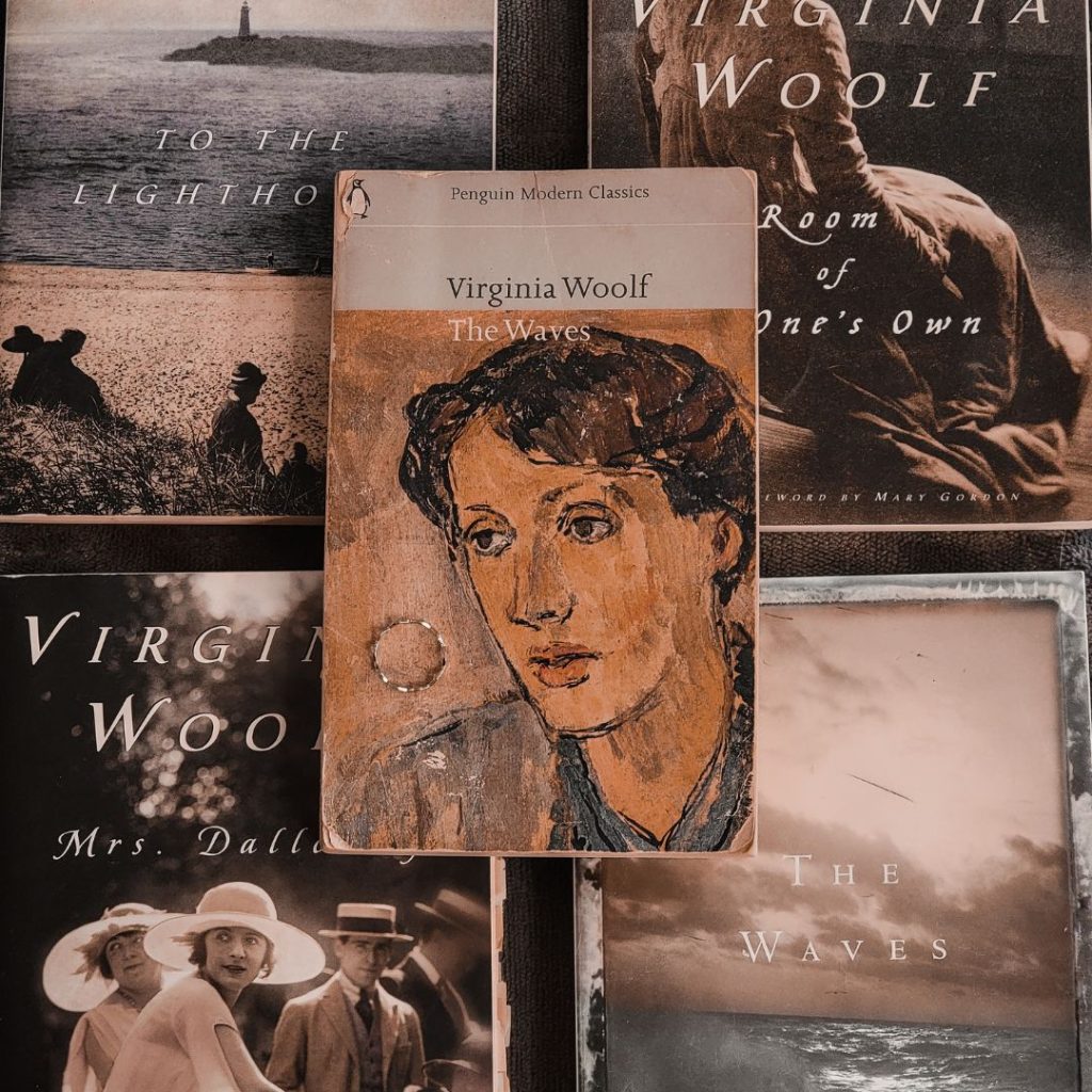 Four books are arranged in a square shape, with on book on top of them in the middle. They are all books by Virginia Woolf. Two of them are beach scenes, one is a group of people, two women and two men, and another is a woman sitting in an old wooden chair. The book on top has an old painting of a person with short brown hair and a blur collared shirt. A silver ring is placed above the person's shoulder.