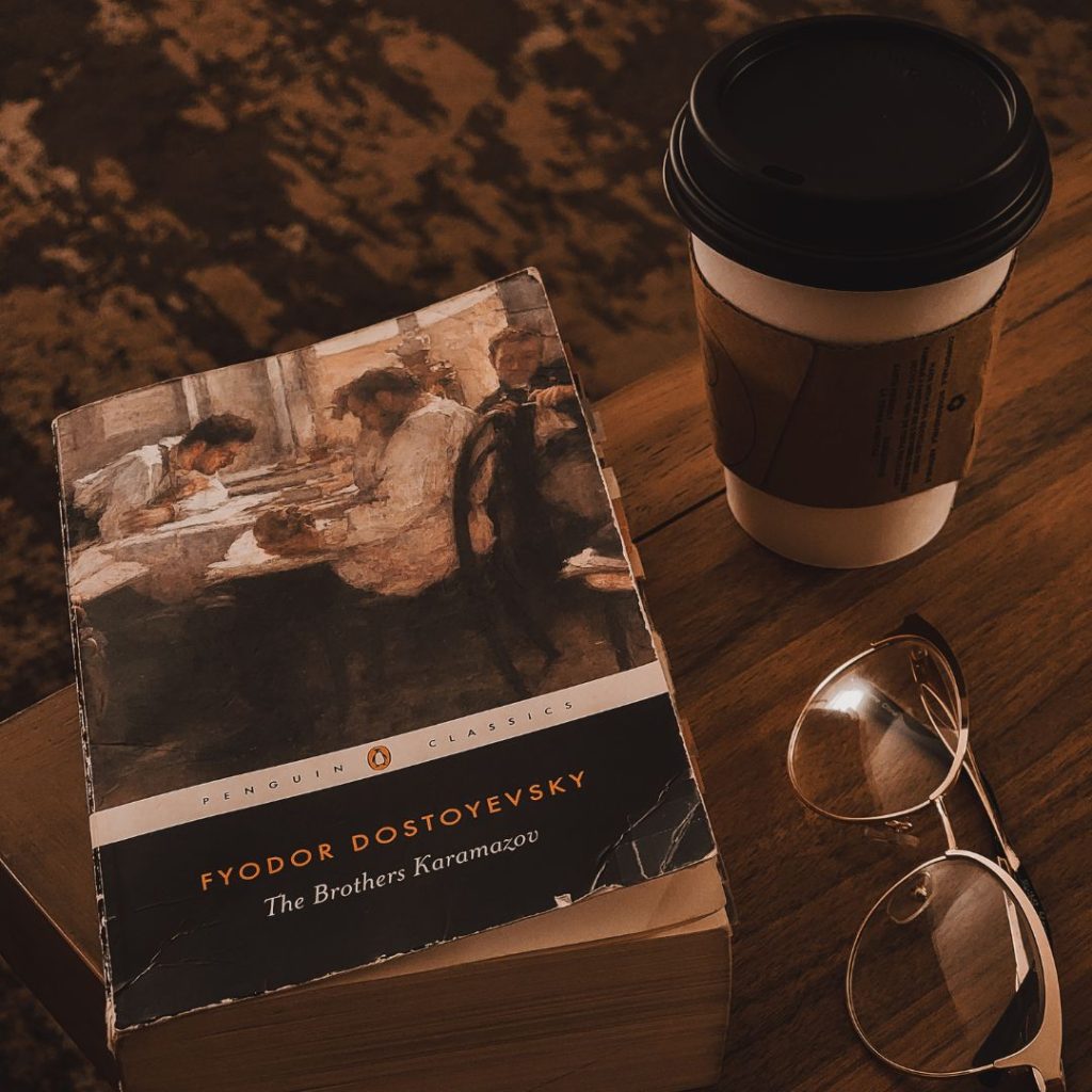 The book The Brothers Karamazov by Fyodor Dostoyevsky sits on the corner of a dark brown wooden table next to a coffee cup and a pair of glasses.
