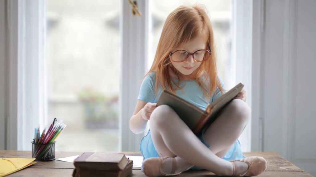 A little girl with glasses reading on a desk.