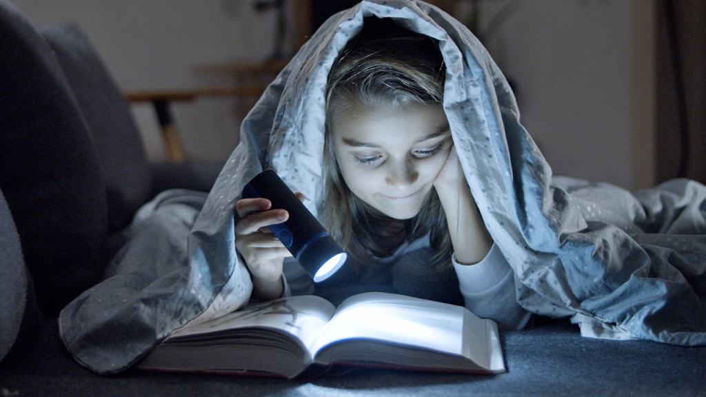 A girl draped in blanket, reading with her flashlight.