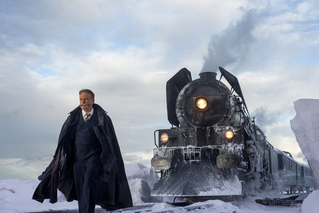 Hercule Poirot standing in front of the Orient Express in a flowing black jacket.