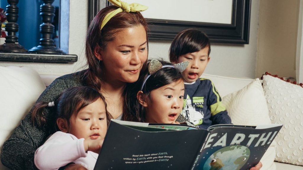 A mom holds a picture book titled "Earth!" while her three young kids are on her lap. The middle kid looks like she is mid sentence, the younger kid on the left is focused on touching the book, and another kid on the right is yawning.