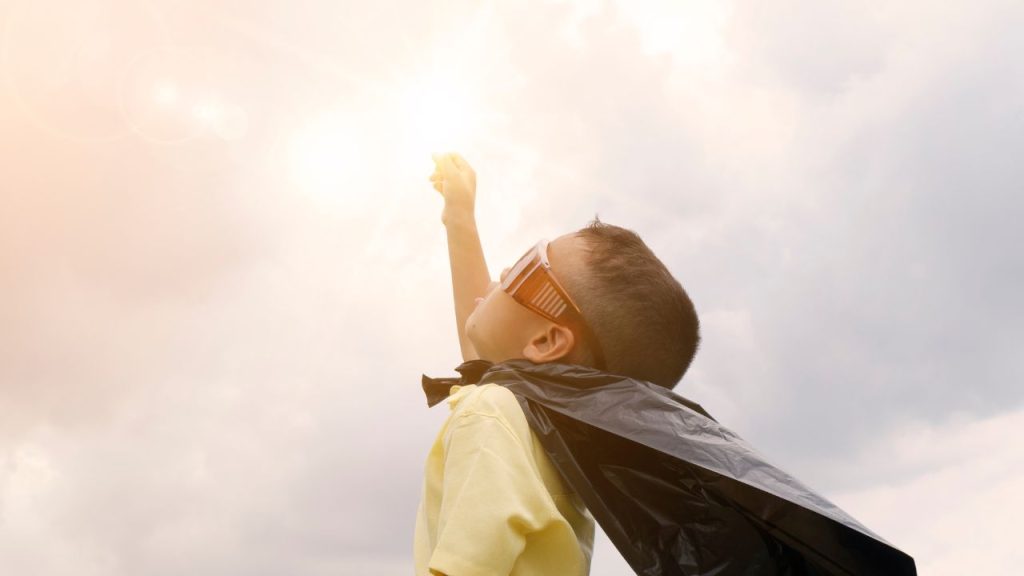 A kid wearing a garbage bag as a cape looks up at a cloudy sky with his mouth slightly open. He is wearing large orange sunglasses.