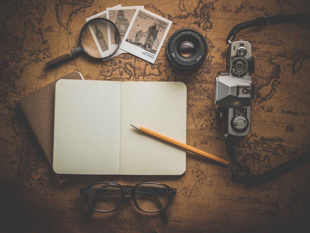 An antique map lays spread out on a table, with an open and a closed notebook placed beneath it. On top of the map are a pencil, a camera, a magnifying glass, three photographs, and a pair of glasses.