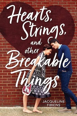 A woman in a black and white dress holds a pair of red converses and leans against a man in a blue cardigan against a brick wall.