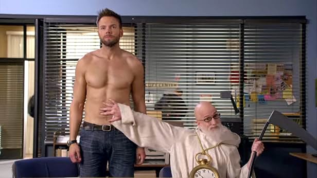 A shirtless man standing with a man dressed as a reaper touches his stomach in a police office.