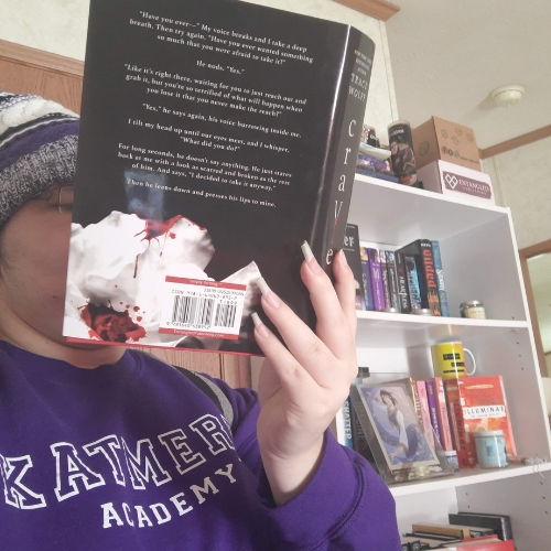 Young woman in a purple crewneck, holding an open book to her face, with a bookshelf in the background.