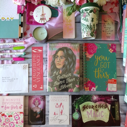 Pink and green spread of stationary, on a wooden tile surface, featuring the book "I'd Rather Burn Than Bloom" by Shannon C.F. Rogers as a centerpiece.