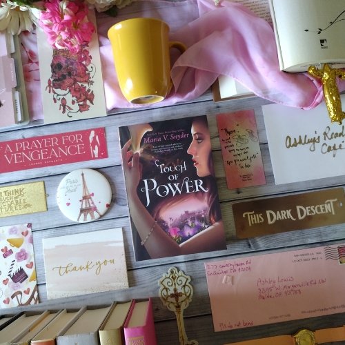 Cover of "Touch of Power" by Maria V. Snyder surrounded by pink and gold stationary, and a yellow mug, on a wooden tile surface.