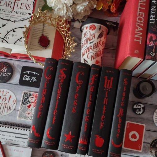 Vertically stacked, hardcover editions of "The Lunar Chronicles" book series by Marissa Meyer, surrounded by different red, black, and white items, and a gold crown, on a wooden tile surface.