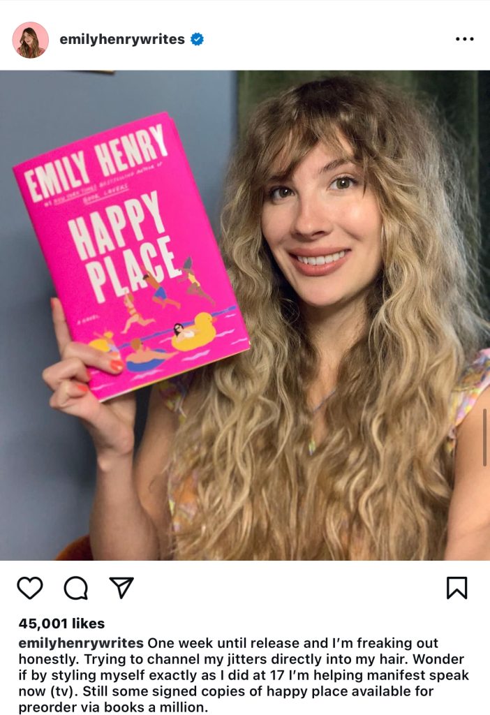 Instagram post by Emily Henry of her holding up her book "Happy Place." She has long blonde hair and the book is bright pink with characters jumping and swimming in a body of water.