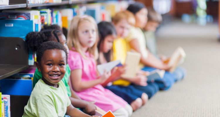 Several young children sitting in a row with their backs against a library bookshelf. The child closest to the camera is looking at the camera and smiling.
