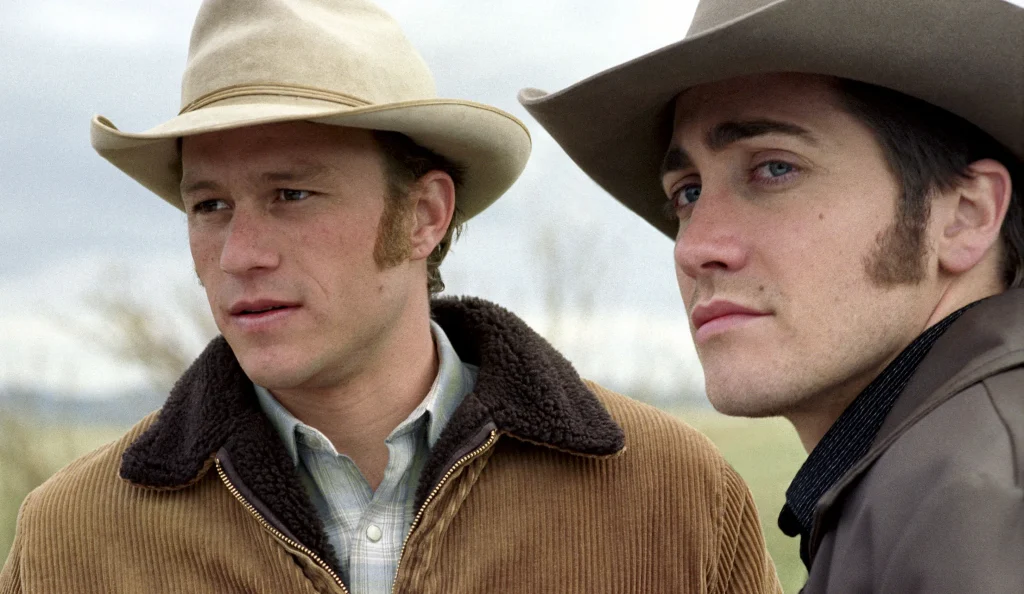Both protagonists are staring intently at something off-camera, with a blurred nature background emphasizing their expressions and emotions. The scene captures their rugged features and deep connection, set against the serene and expansive outdoor backdrop.
