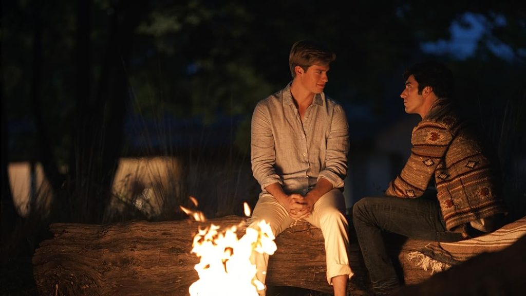 Two men sit outside on logs around a fire at night, facing each other.