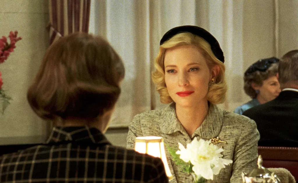 A refined woman with blonde hair, wearing a vintage grey dress and black hat, sits at an elegant table across from another woman whose back is to the camera. She is gazing intently with a slight smile, set against a backdrop of sophisticated decor.