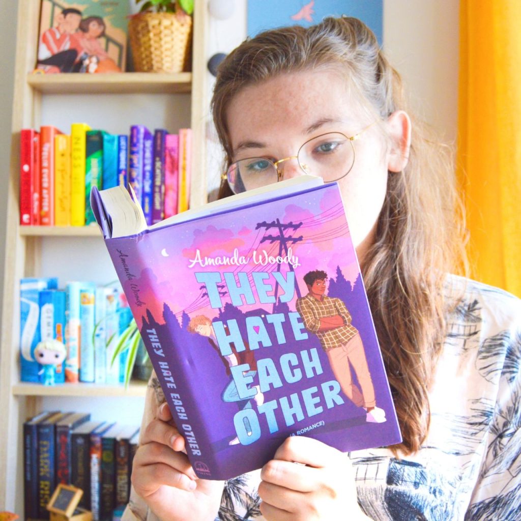 Eloise holding THEY HATE EACH OTHER by Amanda Woody in front of a rainbow color coded bookshelf.