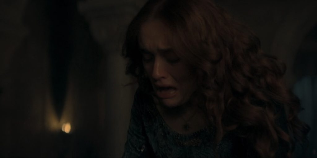 A woman with red hair and a green elaborate gown looks down, crying. We can only see from her mid-chest up.