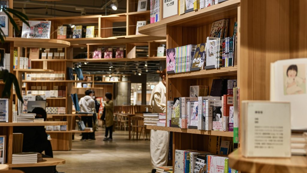 Library of Japanese manga and other Asian publications on beige wooden shelves, and three library goers in the background.