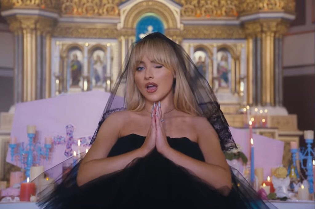 Sabrina Carpenter winking at the camera wearing a black tulle dress and a matching headpiece in front of the alter in a church.
