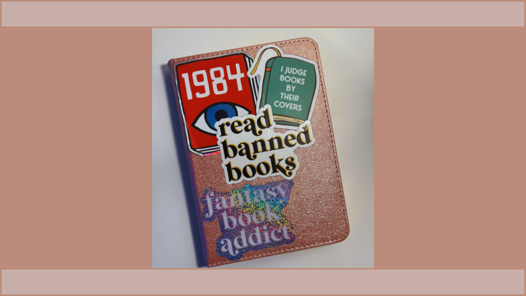 My personal Kindle that has a pink shimmering case with a foldable cover. My stickers include the book 1984, a book with the quote "I Judge Books By Their Covers"," a "Read Banned Books" sticker, and a shimmering "Fantasy Book Addict" sticker.