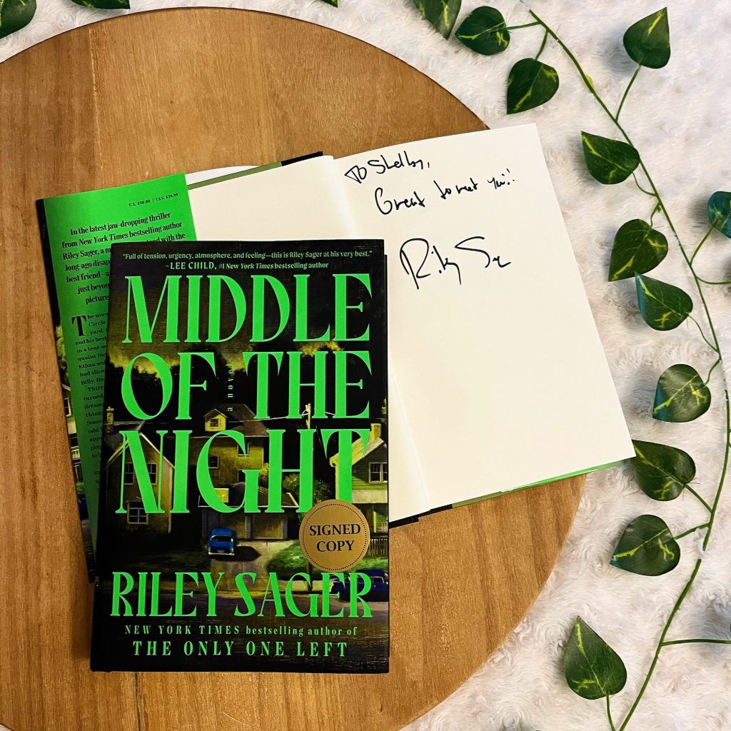 A signed copy of Middle of the Night by Riley Sager on a circular wooden tray surrounded by a vine of green leaves.