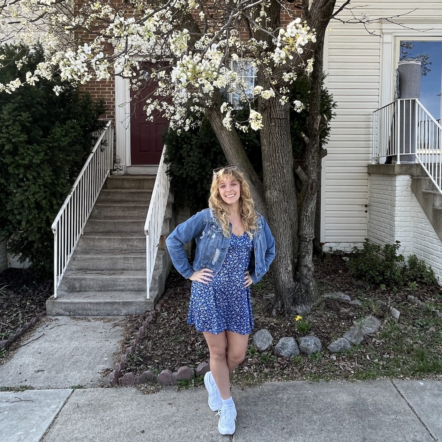 Shelby wearing a floral blue dress and denim jacket and standing in front of a tree blooming white flowers.
